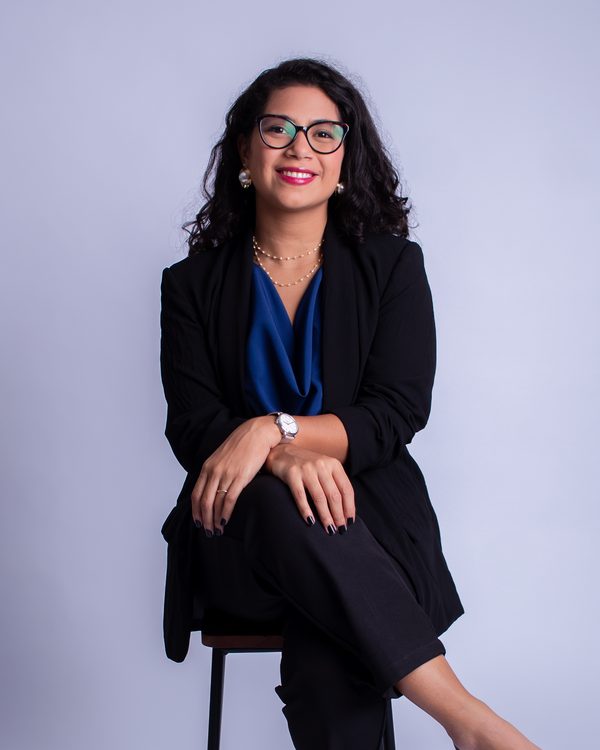 Anniele Costa, de blazer preto, sentada em uma banqueta com as mãos cruzadas sobre o joelho, sorrindo.