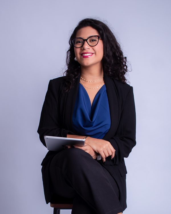Anniele Costa, de blazer preto e blusa azul, sentada em uma banqueta, segurando um tablet e sorrindo.